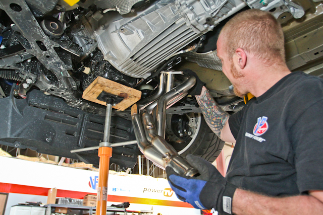 man fitting exhaust headers into a car on lift