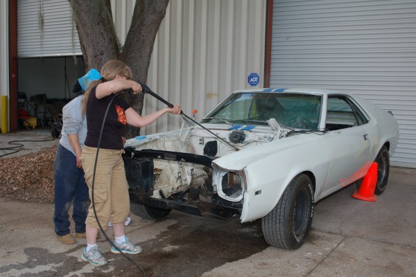 women pressure washing an old AMC AMX muscle car