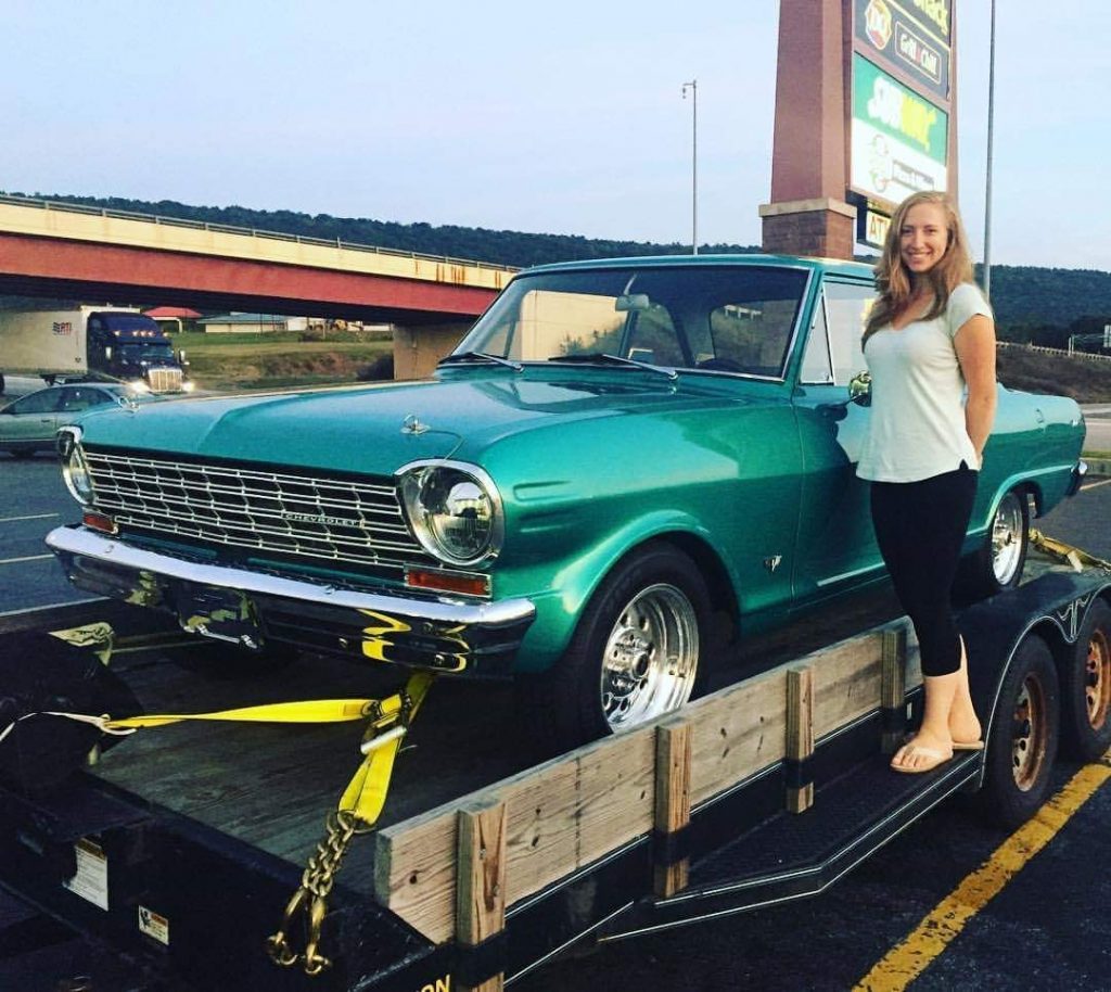 a woman stands next to a chevy 2 nova on a trailer