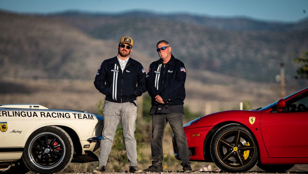 Two men posing near classic cars on a road